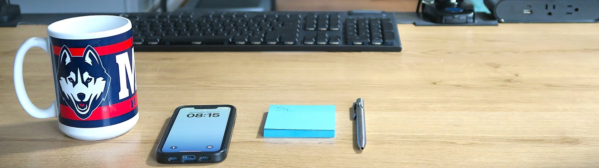 UConn coffee mug, a smart phone showing 8:15am, notepad, and pen on desk with a keyboard in the background